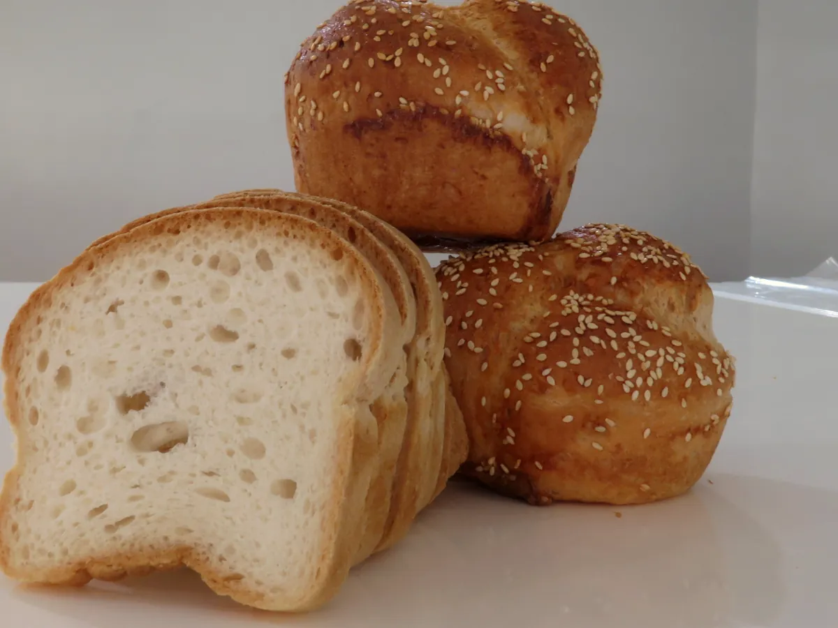 Unstyled photo of sliced gluten free bread showing a soft white crumb, next to two sesame-topped rolls